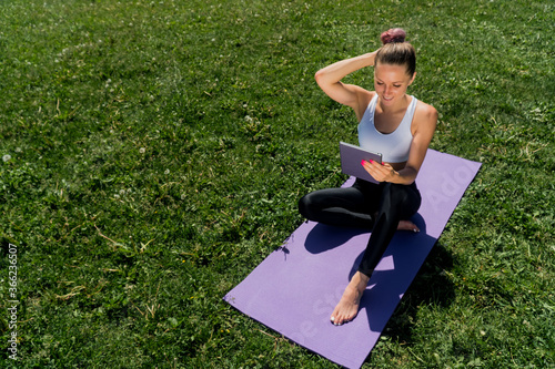 A young girl in sportswear with a beautiful smile, the girl lies on the grass on a yoga mat and looks in a digital tablet.