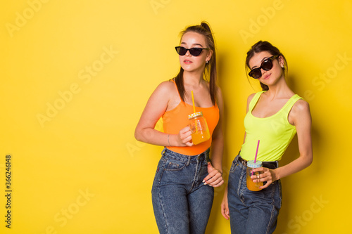 Two cheerful young hipster natural beauty girls posing together in sun glasses on yellow background © Andreshkova Nastya