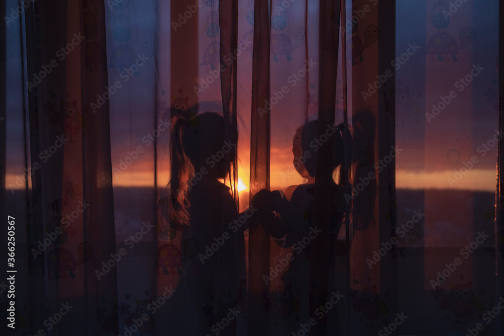 silhouettes of two girls on the windowsill behind the curtain against the sunset sky