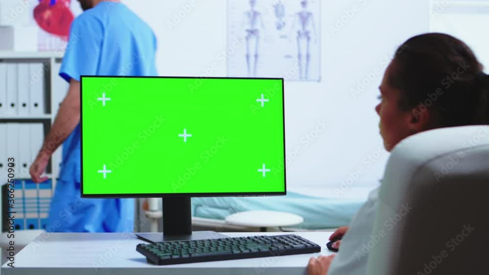 Doctor working on computer with green screen in hospital and assistant looking at x-ray. Desktop with replaceable mockup screen in medical clinic while doctor is checking patient radiography for
