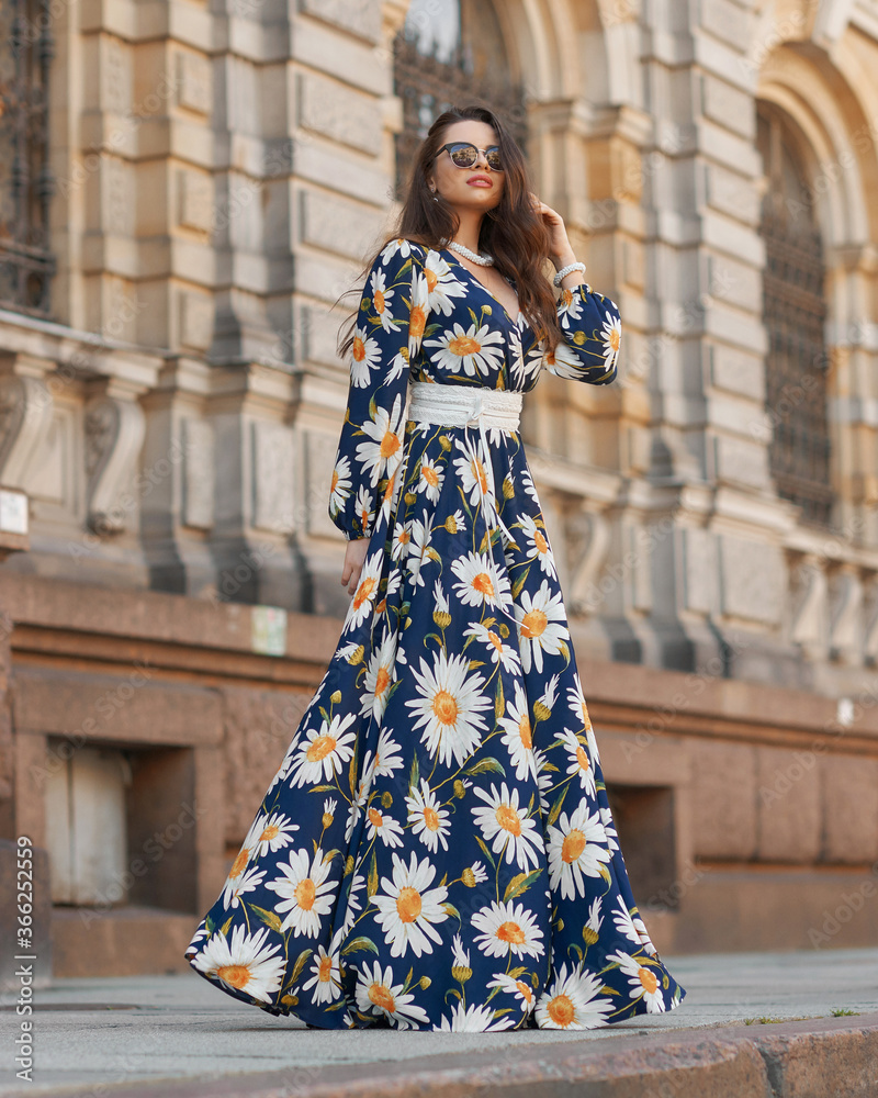 Elegant woman in blue long sundress with floral design posing at city street. Female model wearing dress. Girl with makeup and wavy hair in sunglasses