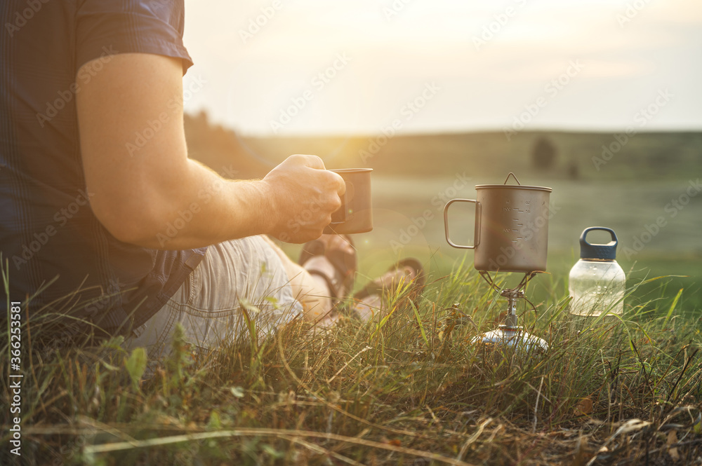 Crop view of man traveler holds a mug of hot tea at sunset. Adventure, travel, tourism and camping concept. Tourist drinking tea from a mug in the camp. Copy space