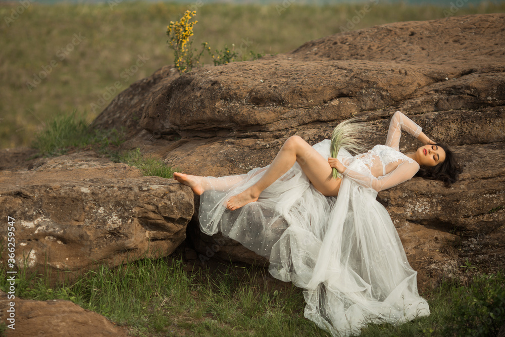 Naklejka premium beautiful young female with makeup and hairstyle in a gentle wedding peignoir lies on the stones outdoors