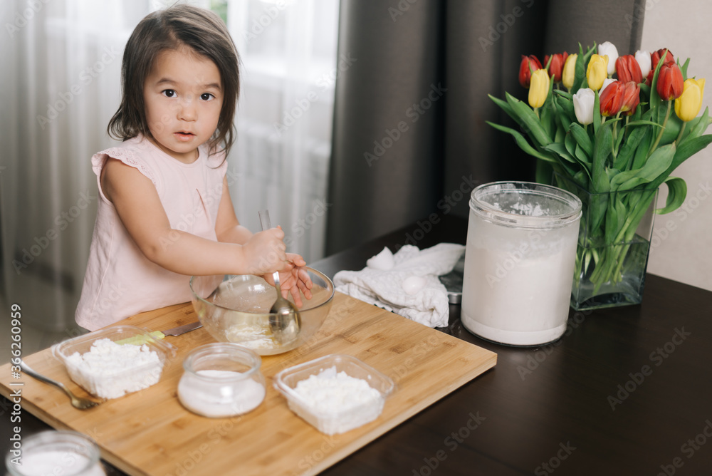 Little two-year-old girl wearing white dress is using stainless spoon to mix the egg for making bakery at kitchen, looking at the camera. Little two-year-old girl wearing white dress is using stainless spoon to mix the egg for making bakery at kitchen, looking at the camera.
