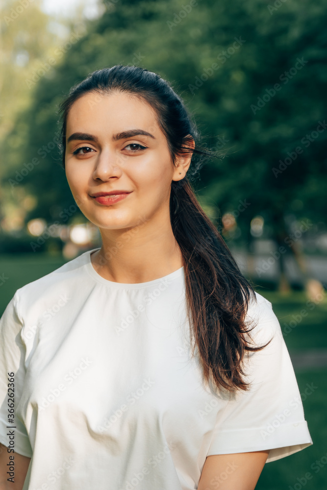 One young beautiful caucasian brunette in a white t-shirt posing in the park on a sunny summer day against the backdrop of green trees. Arabian carefree woman smiling. One young beautiful caucasian brunette in a white t-shirt posing in the park on a sunny summer day against the backdrop of green trees. Arabian carefree woman smiling.