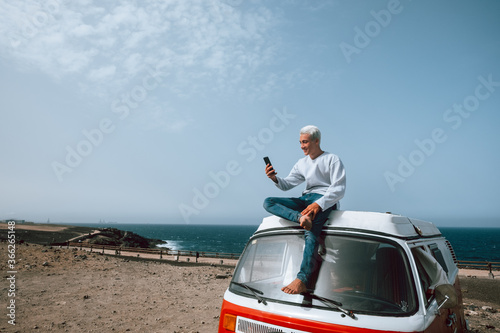 one man alone and isolated sitting on a minivan or camper using his cellphone smiling and looking at it - digital nomad people lifestyle at the beach using technology with the sea at the background