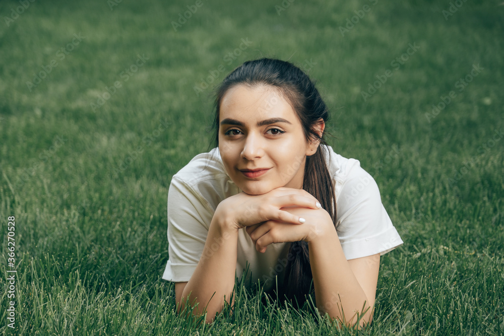 portrait one young beautiful brunette with dark eyes in a white t-shirt lies on the grass holding her head with her hands and looks. Caucasian woman on a summer day. Place for text. portrait one young beautiful brunette with dark eyes in a white t-shirt lies on the grass holding her head with her hands and looks. Caucasian woman on a summer day. Place for text.