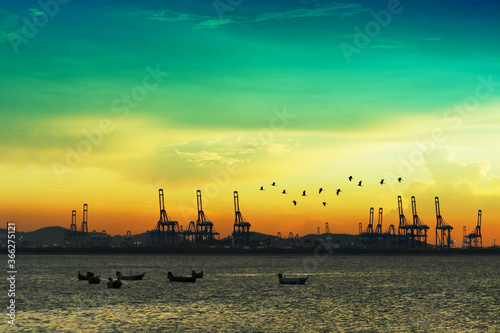 birds flying over cranes on seaport and sunset colorful sky and fishing boats on sea