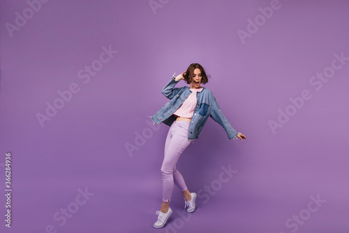 Full-length portrait of attractive slim girl wears white sport shoes. Indoor photo of enchanting caucasian woman dancing on violet background.