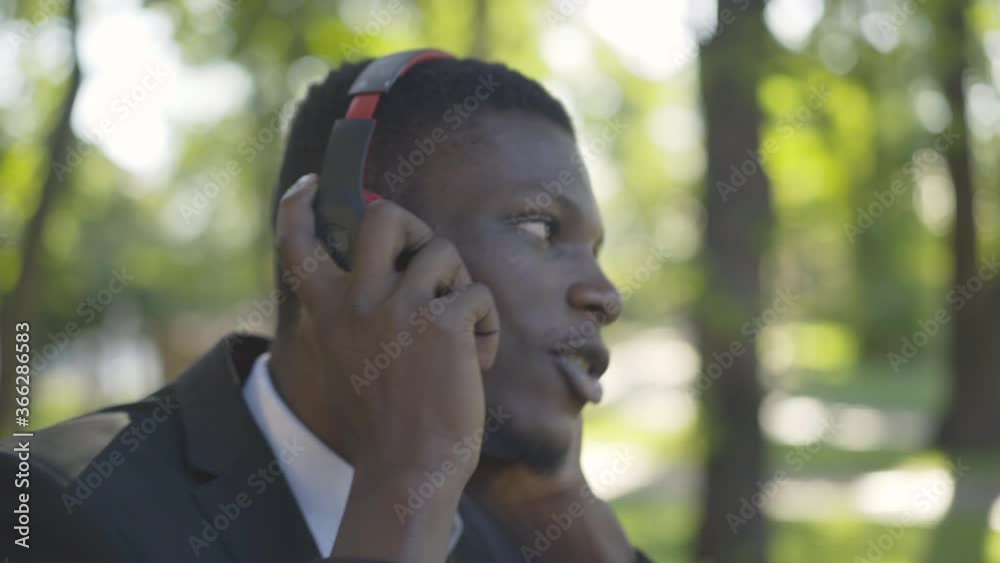 Close-up face of cheerful African American man enjoying music in headphones. Side view portrait of confident businessman in elegant suit singing out loud in sunny summer park. Joy, lifestyle, leisure. Close-up face of cheerful African American man enjoying music in headphones. Side view portrait of confident businessman in elegant suit singing out loud in sunny summer park. Joy, lifestyle, leisure.