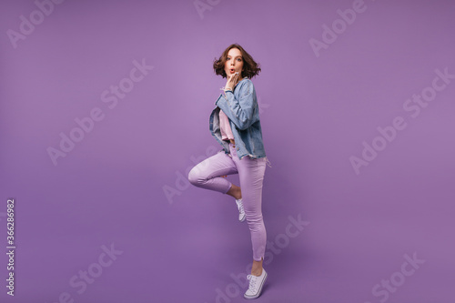 Full-length portrait of funny charming woman dancing in studio. Lovely slim girl in oversize jacket jumping on purple background.