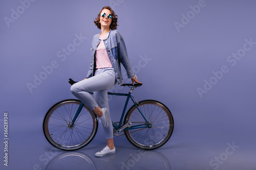 Fototapeta Naklejka Na Ścianę i Meble -  Full-length portrait of laughing glad woman in denim attire. Short-haired girl dancing beside bicycle.