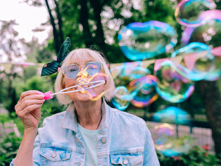 senior (old) stylish woman with gray hair and in blue glasses and jeans jacket blowing bubbles outdoors. Holidays, party, anti age, fun concept
