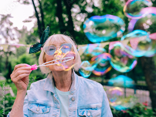 Valokuva senior (old) stylish woman with gray hair and in blue glasses and jeans jacket blowing bubbles outdoors