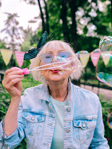 senior (old) stylish woman with gray hair and in blue glasses and jeans jacket blowing bubbles outdoors. Holidays, party, anti age, fun concept
