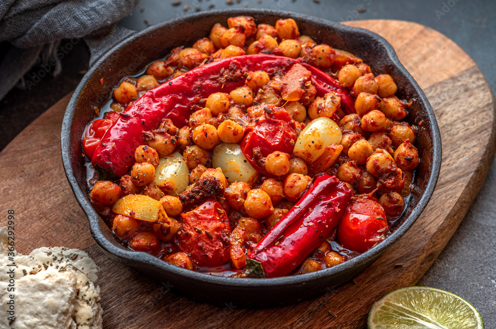 Fototapeta premium Spicy chickpeas with hot red peppers, tomatoes, cumin, garlic and other spices in a frying pan on a black background. Chickpeas stewed with vegetables and spices, oriental cuisine. Vegetarian food.