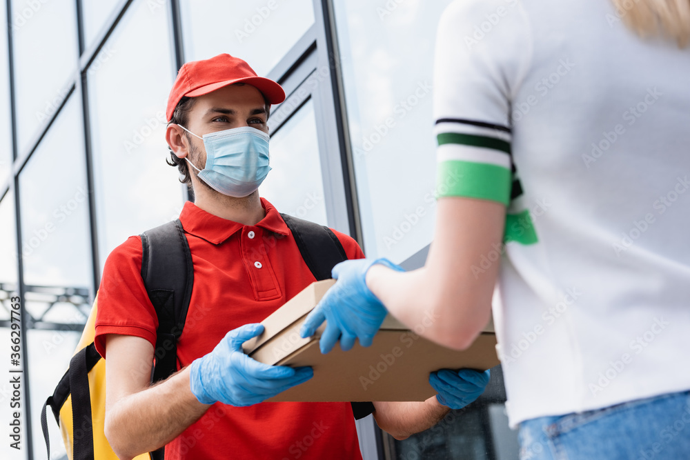 Selective focus of courier in medical mask giving takeaway pizza to girl in latex glove on urban street Selective focus of courier in medical mask giving takeaway pizza to girl in latex glove on urban street