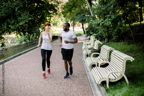 Horizontal shot of young multi racial couple, wearing white t-shirts and black pants, jogging in the city park alley, at early morning with sunrise in background