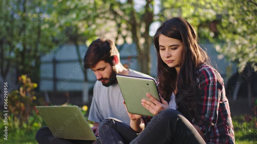 Charismatic woman and guy while sitting on the grass they concentrated working or studying online using the gadgets in a per sunny day Charismatic woman and guy while sitting on the grass they concentrated working or studying online using the gadgets in a per sunny day