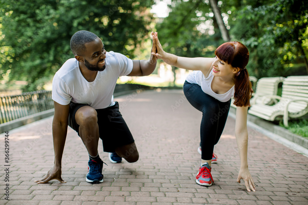 Obraz premium Young fitness couple of African bearded man and attractive Caucasian woman, ready to start running at the park, giving high five each other and smiling. Jogging together outdoors