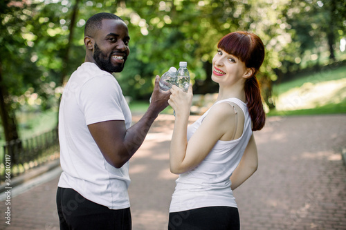 Fototapeta Naklejka Na Ścianę i Meble -  Sport, fitness, jogging and lifestyle concept. Cheerful smiling multiethnical couple with bottles of water, posing to camera during their break after running workout outdoors at the city park
