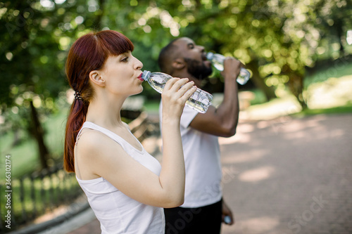 Water break, sport and fitness concept. Happy young multiethnic friends, Caucasian woman and African man, drinking water after jogging in park. Side view, focus on woman face