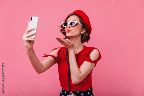 Stunning french woman sending air kiss while taking picture of herself. Indoor portrait of romantic curly lady in betet making selfie on pink background.