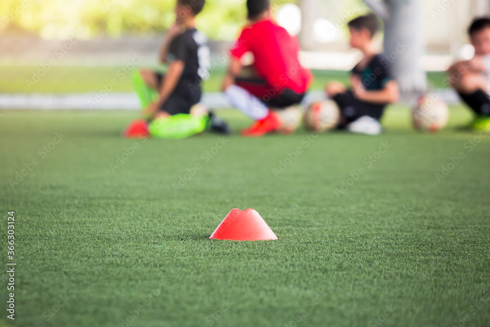 selective focus to red cone marker is soccer training equipment on green artificial turf with blurry kid players training background. Material for trainning class of football academy. selective focus to red cone marker is soccer training equipment on green artificial turf with blurry kid players training background. Material for trainning class of football academy.