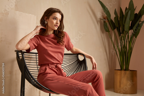 Beautiful brunette woman natural makeup wear fashion clothes casual dress code office style total pink blouse and pants suit, romantic date business meeting armchair interior stairs flowerpot. photo