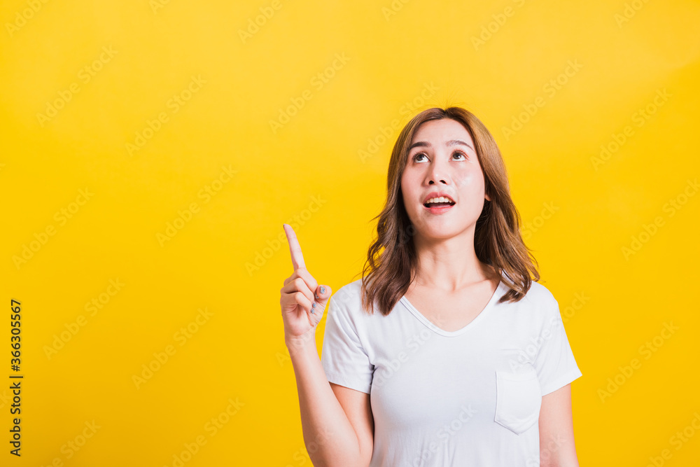 Asian Thai happy portrait beautiful cute young woman standing wear white t-shirt makes gesture two fingers point upwards above looking above, studio shot isolated on yellow background with copy space Asian Thai happy portrait beautiful cute young woman standing wear white t-shirt makes gesture two fingers point upwards above looking above, studio shot isolated on yellow background with copy space