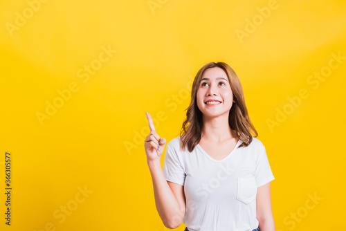 Asian Thai happy portrait beautiful cute young woman standing wear white t-shirt makes gesture two fingers point upwards above looking above  studio shot isolated on yellow background with copy space