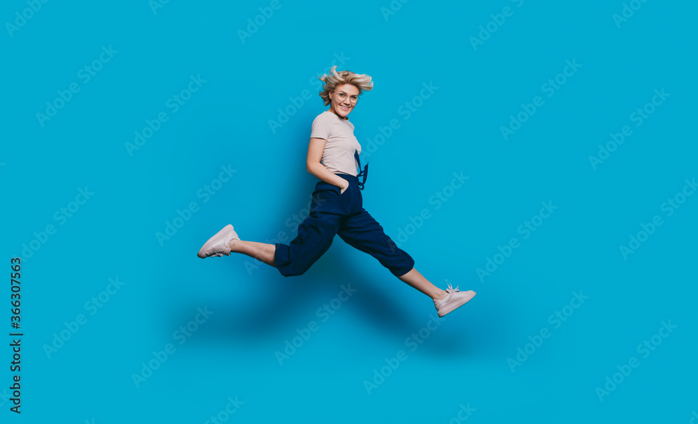 Energetic charming woman with blonde hair smiling at camera and jumping on a blue studio wall