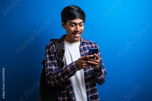 Young asian student smile and use phone isolated on blue background. The guy looks happy chatting with his friend