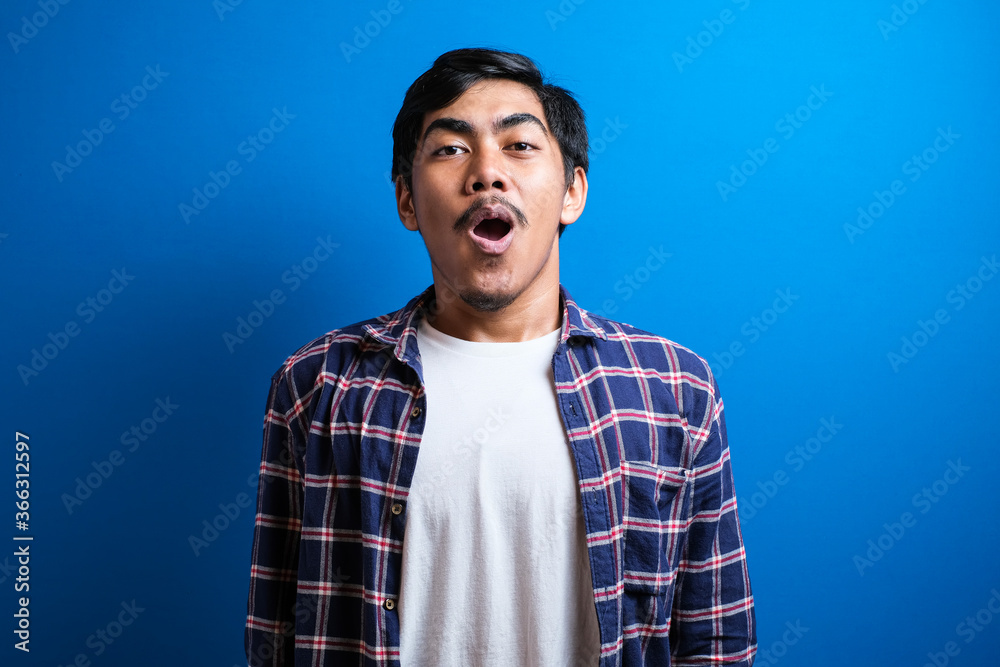Shocked Asian man dressed in casual ith open mouth and hands up looks to the camera. Surprised young asian student guy on blue studio background