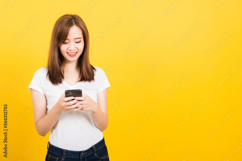 Asian happy portrait beautiful cute young woman teen smile standing playing game or writing message on smartphone looking to the phone isolated, studio shot on yellow background with copy space Asian happy portrait beautiful cute young woman teen smile standing playing game or writing message on smartphone looking to the phone isolated, studio shot on yellow background with copy space