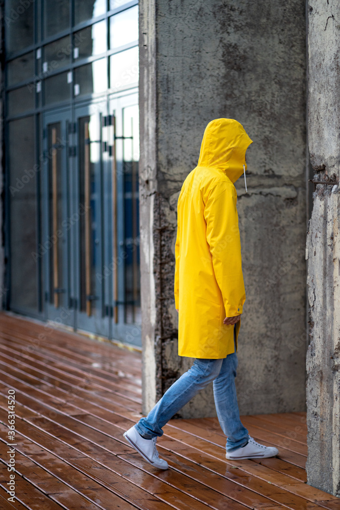 Man in a yellow raincoat with the hood on walks in rainy weather near the building on wet wooden floor, side view. Outdoor. Man in a yellow raincoat with the hood on walks in rainy weather near the building on wet wooden floor, side view. Outdoor.