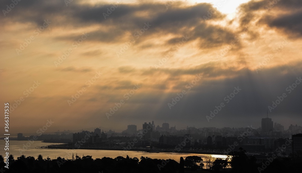 Fototapeta premium Sunset landscape on the city coast. Clouds and sun rays over the buildings.
