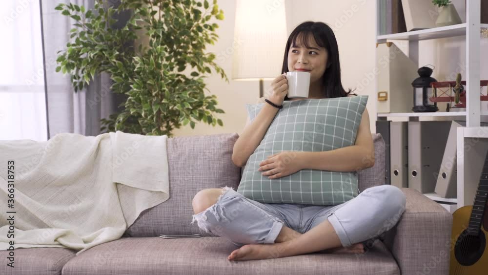lovely asian lady having tea in a relaxing mood on living room couch. Young lady in love holding cushion beamed with happiness thinking of her thoughtful boyfriend. lovely asian lady having tea in a relaxing mood on living room couch. Young lady in love holding cushion beamed with happiness thinking of her thoughtful boyfriend.