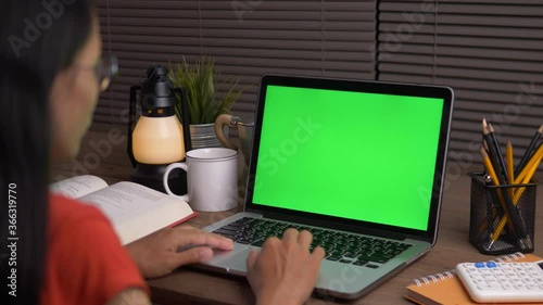 Over the shoulder shot of a business woman working in office interior on pc on desk, looking at green screen. Office person using laptop computer with laptop green screen, Dark Office.