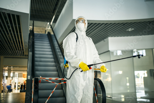 Portrait of a man in a sanitizing disifection suit holding spray near the escalator in an empty shopping mall. A volunteer cleaning up the public places to prevent covid-19. Health awareness concept.