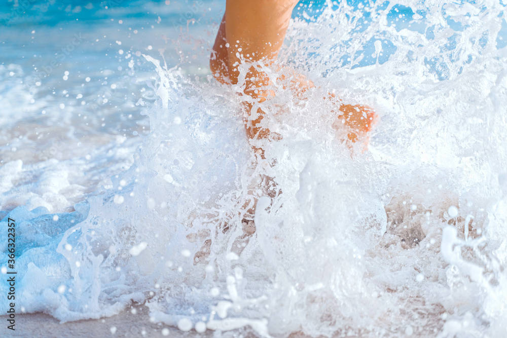 A wave breaks on the shore and the beautiful legs of a young girl. Ocean foam and sea water splashes. Beach holidays, travel concept.