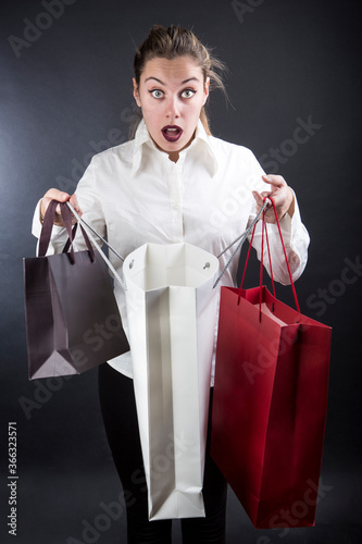 ragazza bionda con i capelli raccolti e una camicia bianca guarda stupita dentro con le buste degli acquisti in mano, isolata su sfondo nero photo