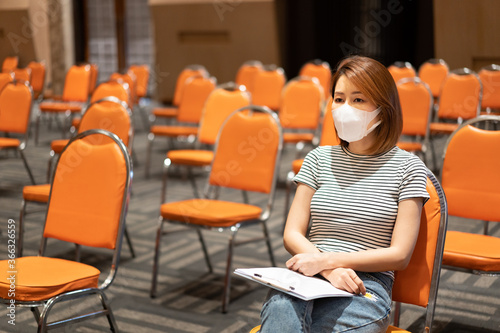 An Asian woman with a golden-bronze hair wearing a striped shirt wear facemark sitting and waiting for something in a chair amid an empty chair in orange. photo