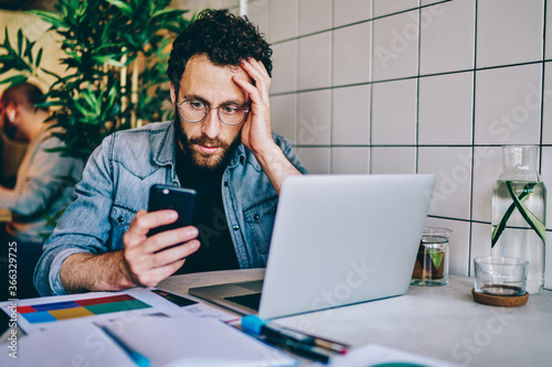 Worried bearded young man reading sms message with bad notification on modern smartphone sitting at laptop computer in cafe.Frustrated and upset hipster guy watching shocked video in blog on cellular