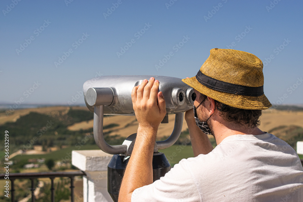 Photo of a young and attractive man wearing a reusable facce mask looking to the nature through some binoculars in a view point. Tourism during coronavirus outbreak