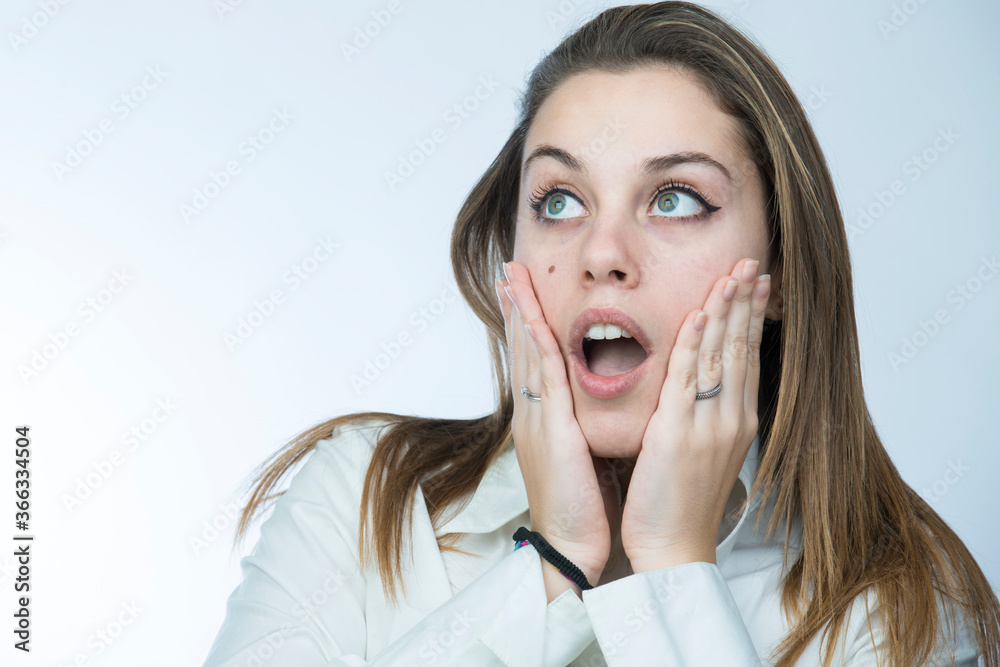 ragazza bionda con camicia bianca si porta le mani alla faccia e guarda lontano con aria meravigliata, isolata su sfondo chiaro ragazza bionda con camicia bianca si porta le mani alla faccia e guarda lontano con aria meravigliata, isolata su sfondo chiaro