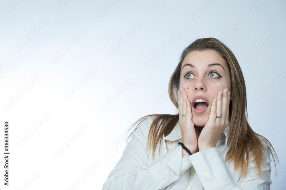 ragazza bionda con camicia bianca si porta le mani alla faccia e guarda lontano con aria meravigliata, isolata su sfondo chiaro ragazza bionda con camicia bianca si porta le mani alla faccia e guarda lontano con aria meravigliata, isolata su sfondo chiaro