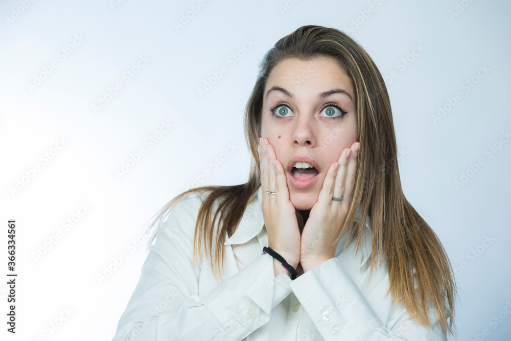 ragazza bionda con camicia bianca si porta le mani alla faccia e guarda lontano con aria meravigliata, isolata su sfondo chiaro