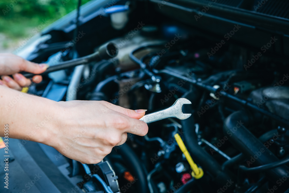 Woman is repairing broken car. Accident situation while travelling. Opened bonnet of vehicle. Girl is holding instruments for fixing.