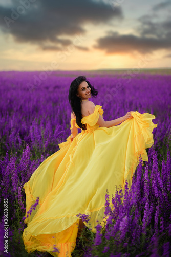 Summer mood. A woman in a luxurious yellow dress walks along a purple blooming field with her back to the camera.